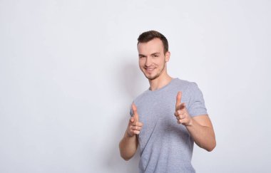 Gri tişörtlü genç bir adam gülümsüyor, beyaz bir arka planda izole edilmiş olumlu duygular ve mutlulukla baş parmaklarını gösteriyor. Kalite ve tavsiye ile tatmin olmak. Boşluğu kopyala.