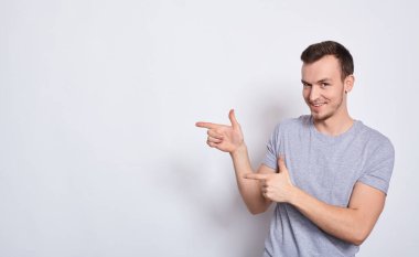 Yakından, gri tişörtlü, genç, yakışıklı, atletik beyaz adam, gülümsüyor, düz görünüyor, parmaklarını sola doğru gösteriyor, beyaz bir arka planda, metnin için kopyalama alanı ile izole edilmiş. Boşluğu kopyala.