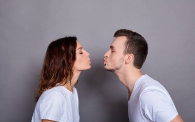 Beyaz tişörtlü, beyaz erkek ve kadınlar, gözlerini kapatmış, öpüşüyor, gri bir arka planda stüdyoda poz veriyorlar..