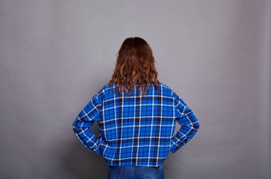 Kafeste mavi gömlekli, kahverengi uzun saçlı, genç beyaz bir kızın görüntüsü, ellerini kalçasına koyar, sırtını kameraya çevirir, gri bir arkaplana karşı poz verir..
