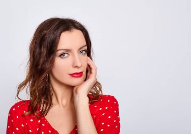 Kırmızı rujlu, parlak akşam makyajlı, güzel, beyaz bir kızın portresi, kırmızı bir elbise giyiyor, yüzüne dokunuyor, beyaz bir arka planda poz veriyor. Boşluğu kopyala.