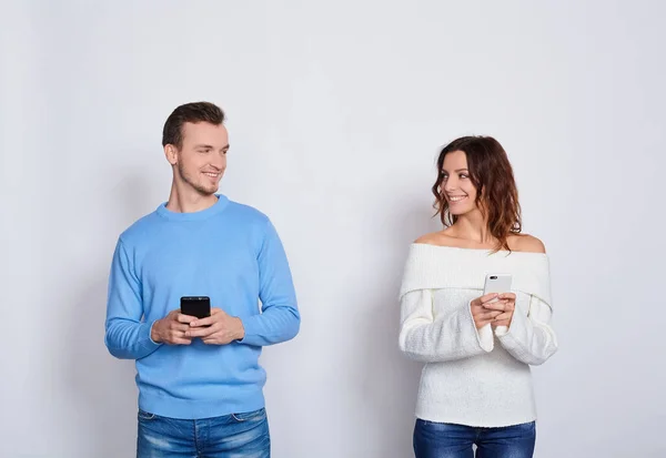 照片上 积极镇定的男人和女人穿着休闲装 面带微笑 相互凝视 同时使用白色背景的手机 — 图库照片
