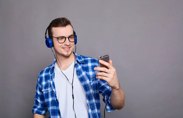 一个年轻可爱的家伙 穿着蓝色衬衫和白色T恤 穿着灰色背景的衣服 拿着耳机听音乐 复制空间 — 图库照片