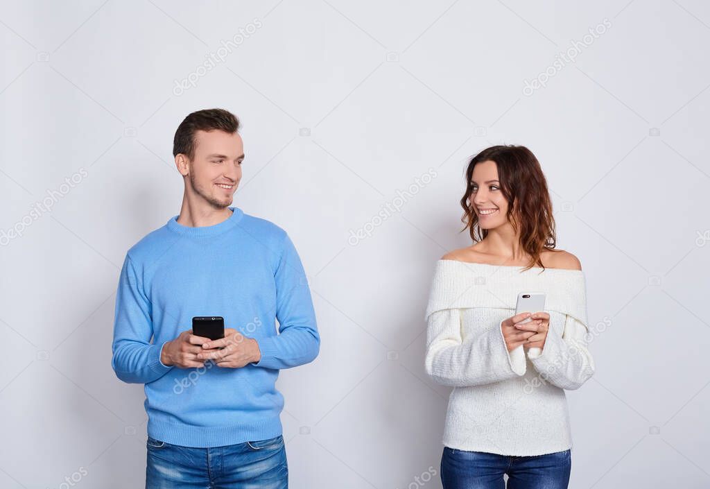 Photo of positive calm people man and woman in casual light clothes smiling, looking at each other, while both using mobile phones isolated on a white background.