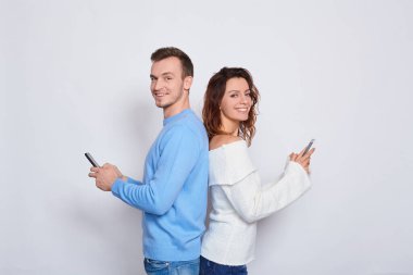 Beyaz kazaklı güzel, tatlı, hassas bir kadın, mavi kazaklı yakışıklı bir adam, sırt sırta verin ve akıllı telefonlarda 3G internet üzerinden SMS yazın, kameraya bakın ve beyaz bir arka planda gülümseyin..