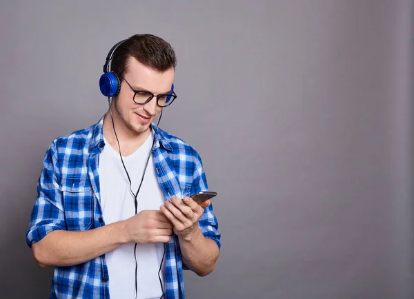 一个穿着白色T恤 蓝色格子花衬衫的年轻英俊白人小伙子 用耳机听音乐 阅读短信 写短信 用智能手机 在灰色背景上摆姿势 复制空间 — 图库照片