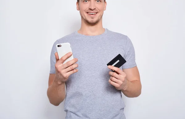 一个穿着灰色T恤衫的英俊年轻人 被裁剪成这样 他正在考虑在网上购物 手里拿着一张信用卡和一部白色背景的智能手机 — 图库照片