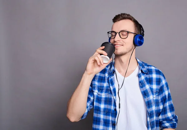 A young happy cheerful joyful man in glasses, in a white T-shirt and blue shirt, holds a black coffee cup, listens to his favorite music with headphones, closed his eyes and smiles with pleasure.