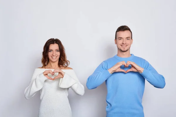 Funny cheerful happy young Caucasian lovers in light fashionable clothes, stand next to each other, show their hands heart, are in love, test emotions, smile on a white background.