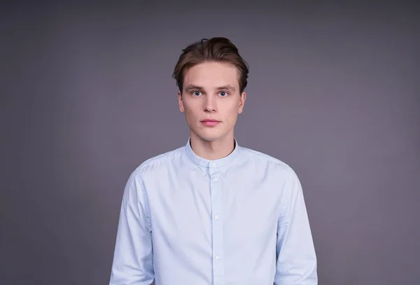 Cute cute young Caucasian guy with blond hair and blue eyes, stands straight, confident and calm looking into the camera, posing on a gray background in the studio.