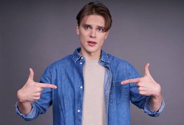 Sarı saçlı, mavi gözlü, bej tişörtlü ve kot gömlekli yakışıklı beyaz adam düz görünüyor, kendini işaret ediyor. Yaşam tarzı kavramı, gençlik..