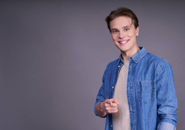 Gri arka planda izole edilmiş genç bir adamın portresi. Sağ elin parmaklarıyla izleyiciyi işaret eden kameraya bakarken gülümsüyor ve mutlu hissediyor. Boşluğu kopyala.