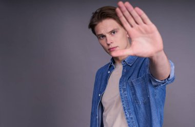 Kes şunu! Hayır, istemiyorum. İstemeyerek kot gömlekli yakışıklı bir adam, reddedilme işareti olarak sol elini uzatmış, inkar jesti, gri arka planda izole edilmiş. Boşluğu kopyala.