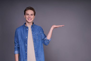 Gri arka planda izole edilmiş, kot gömlek giyen ve sağ tarafta bir şey gösteren, telif alanı da dahil, pozitif gülümseyen genç bir erkeğin yatay portresi. Boşluğu kopyala.