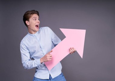 Mavi tişörtlü ve sıradan kotlu genç, şaşırtıcı bir şekilde ağzını açtı, elinde büyük pembe bir ok tutuyor, sağ tarafa doğru, gri bir arka planda poz veriyor..