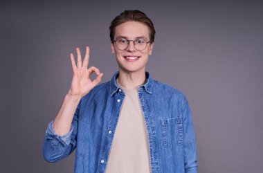 Mavi kot ceketli genç adam mutlu görünüyor, gülümsüyor, el kol hareketi yapıyor, tamam işareti gösteriyor. Beyaz adam parmaklarıyla iyi hareket ediyor, sağ eliyle gri arka planda işaret gösteriyor..