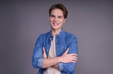 Kolları çapraz duran, geniş kar beyazı bir tebessümle, gri bir arka planda izole bir şekilde gülümseyen, mavi kot gömlekli çekici bir genç adamın portresi..