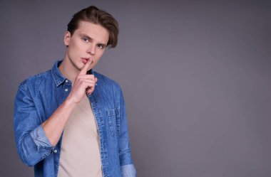 Şşş jest. Gri arka planda, bej renkli bir tişört ve kot gömlek giymiş, dudaklarına parmak basan, sessiz olmasını isteyen, yakışıklı, ciddi, beyaz bir adam. Boşluğu kopyala.