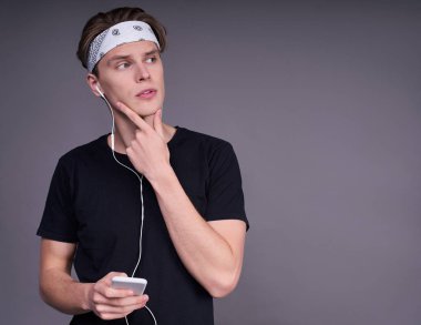 Siyahi tişörtlü, kulaklık takan, telefonu elinde tutan, çenesine eliyle dokunan, sağına bakan, gri bir arka planda duran düşünceli, genç, beyaz bir çocuk..