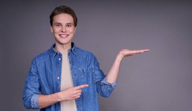 İzole edilmiş gri arka planda gözlük takan yakışıklı Avrupalı model kameraya hayret eder ve gülümserken eliyle poz verir ve sağını işaret ederdi. Boşluğu kopyala.