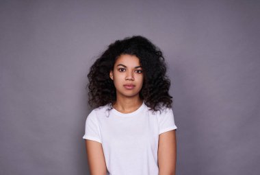 Gri bir arka planda mutlu ve tasasız bir şekilde beyaz dişlerini kameraya gösteren tüylü saçlı, esmer, güzel bir kadının kapalı bir portresi..