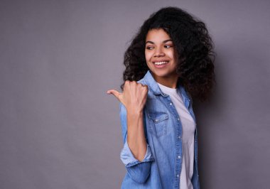 Çekici, sevimli, koyu tenli bir kız, kıvırcık saçlı, beyaz tişörtlü ve kot gömlekli bir öğrenci, parmağını sola çeviriyor, yana bakıyor ve geniş bir şekilde gülümsüyor..