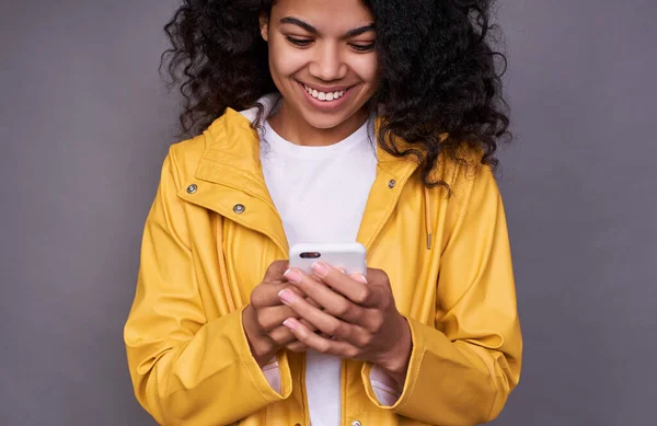 一个快乐的年轻非洲女孩 有着浓密的波浪式发型 身穿黄色雨衣 用手机写下了她心爱的人的短信 现代技术概念 — 图库照片