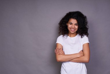 Neşeli bir Afrikalı Amerikalı, hoş bir gülümsemesi, kıvırcık siyah saçları, sağlıklı bir teni var, beyaz bir tişört giyiyor, gri bir arka planda duruyor, iyi bir ruh hali içinde, kollarını göğsünün üzerinde kavuşturuyor..