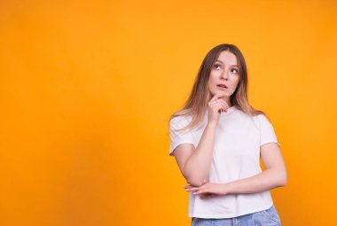 Beyaz tişörtlü genç ve güzel bir kız sarı izole bir arka planda, düşünceli, reklamınız veya mesajınız için uygun yeri düşünüyor..