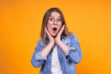 Sersemlemiş Kafkasyalı genç bir kadın gözlerini açıyor, şaşkınlıktan boğuluyor, şok edici habere inanamıyorum, şaşkın, kot gömlek ve yuvarlak gözlük giyiyor..