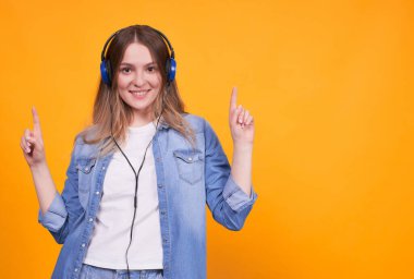 Kulaklıkla müzik dinlemekten hoşlanan, işaret parmaklarını gösteren, sevinen, dans eden, sarı arka planda eğlenen beyaz sarışın bir kızın portresi. Bayan öğrenci kopyalama alanı.