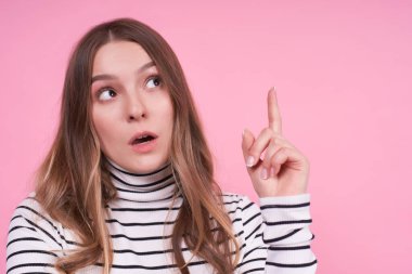 Duygusal genç ve düşünceli sarışın bayan sağa, yakından bakar, etkilenmiş bir şeyler hisseder, işaret parmağıyla işaret eder, pembe arka planda izole edilmiş sıradan bir çizgili süveter giyer, boşluğu kopyalardı..