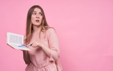 Genç beyaz bir bayan elinde bir kitap tutuyor ve onu ağzında mavi bir kalemle işaret ediyor, hata yaptığı için utanıyor, aynı arka plan, pembe ceket, yanlış anlaşılma ifadesi, sessizlikte korku. Boşluğu kopyala..