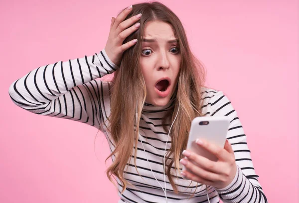 Young emotional Caucasian girl with a shocked surprised excited face with open mouth, clutching her head, looking at the phone, unhappy and not understanding what to do on a pink background.