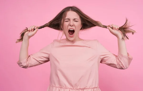 stock image Young Caucasian girl with an open mouth and closed eyes, screams, yawns, holds stretches her hair with her hands in different directions, fooling around with her hair, in a pink background.