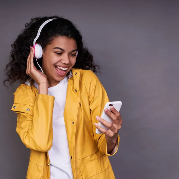 Charming happy young African American girl in white T-shirt and a yellow raincoat, with curly hair, listens to music on headphones, holds a smartphone, listens to her favorite music through the phone.