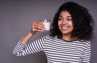 Göz kamaştırıcı bir gülümsemesi olan, koyu tenli tatlı bir kız elinde bir bardak süt tutuyor, ona bakıyor, gri bir arka planda poz veriyor. Kalsiyum, vitamin, diş sağlığı, güçlü kemikler..
