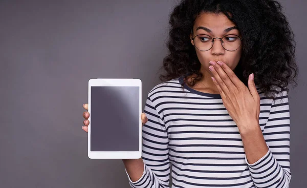 A shocked, amazed, amazed young dark-skinned woman in a striped sweater and glasses is holding a tablet in her right hand and covering her mouth with her left hand, she looks to the side.