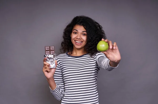 Cute African woman in a striped sweater, with a lush curly hairstyle, holds a green apple and a chocolate bar in her hand, looks straight with smile and shows apple on frame.