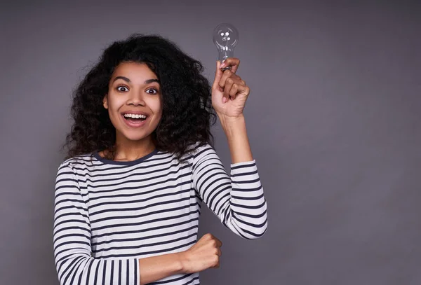 Happy cute young funny african american girl with curly gorgeous hair, in a striped casual jacket, holds a light bulb in her left hand, opens her eyes wide, she has an idea.