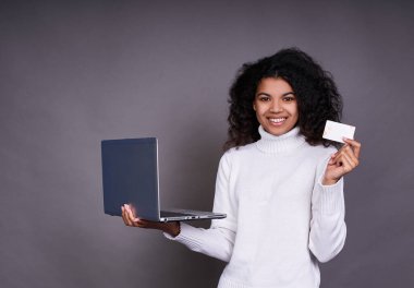 Beyaz kazaklı, dizüstü bilgisayarı ve beyaz banka kartı olan gri stüdyo arka planda izole edilmiş tatlı Afrikalı Amerikalı kız. Mutlu etnik ırk. Kadın internetten alışveriş yapmak için kredi kartı kullanıyor. Boşluğu kopyala.