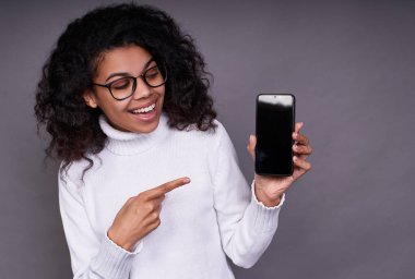 Yakın plan, kıvırcık saçlı, beyaz kazaklı ve gözlüklü neşeli bir Afro-Amerikalı kız elinde bir telefon tutuyor ve sağ elinin parmaklarıyla telefona bakıyor..