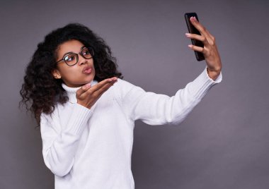 Gözlüklü, siyah tenli ve beyaz kazaklı hoş bir kadın, gri arka planda telefonla selfie çekiyor, kameraya hava öpücüğü gönderiyor..