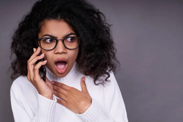 Close-up, shocked surprised young dark-skinned young girl in glasses and a white sweater, with curly hair, speaks on the phone on a gray background, opened her mouth and looks to the side.