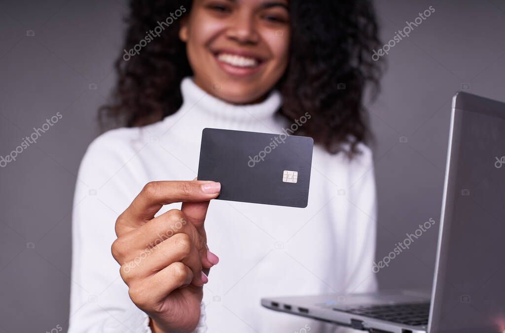 Close-up, smiling young dark-skinned girl in a white sweater, with a laptop, holds a black bank card in her left hand. Concept of online shopping, quarantine purchases.