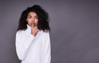 Koyu tenli, beyaz kazaklı, muhteşem kıvırcık saçlı, korkmuş genç bir kız parmağını ağzına dayayıp sessizlik istiyor, o da sırrı öğrendi. Gri bir arka planda fotoğraf. Boşluğu kopyala.