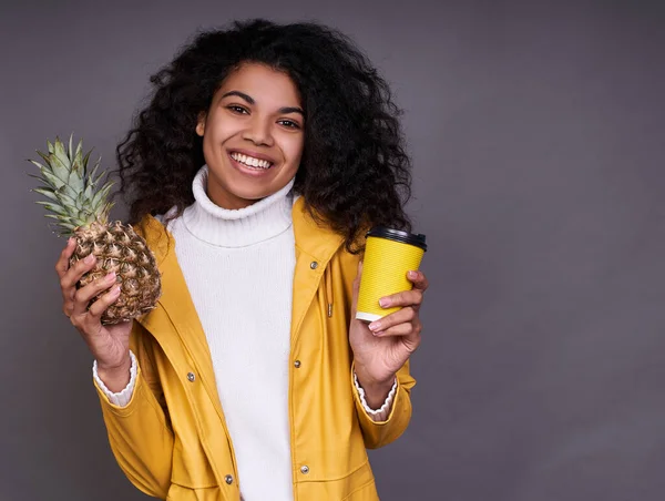 Close-up portrait of a charming dark-skinned young woman with curly hair, in a white sweater and yellow jacket, holding a pineapple and a yellow coffee cup in her hands, looking directly with a smile.