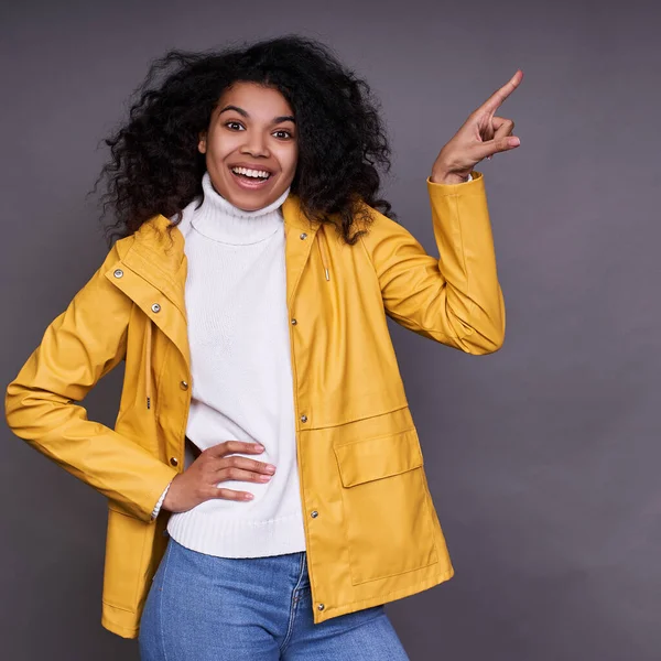 The vertical shot was liked by a dark-skinned woman with an afro haircut pointing to the side with her left hand, smiling and looking straight, isolated on a gray background.