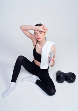 Siyah bluzlu, beyaz bir arka planda oturan, spor yaptıktan sonra elini alnına dayayıp dinlenen ince, beyaz bir kadının üst görüntüsü..