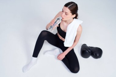 Üst manzara, atletik beyaz bir kız, siyah spor bluzu ve taytlı, şişeden su içiyor, idmandan sonra dinleniyor, boks eldivenleri yakınlarda, beyaz bir arka planda izole bir şekilde yatıyor..
